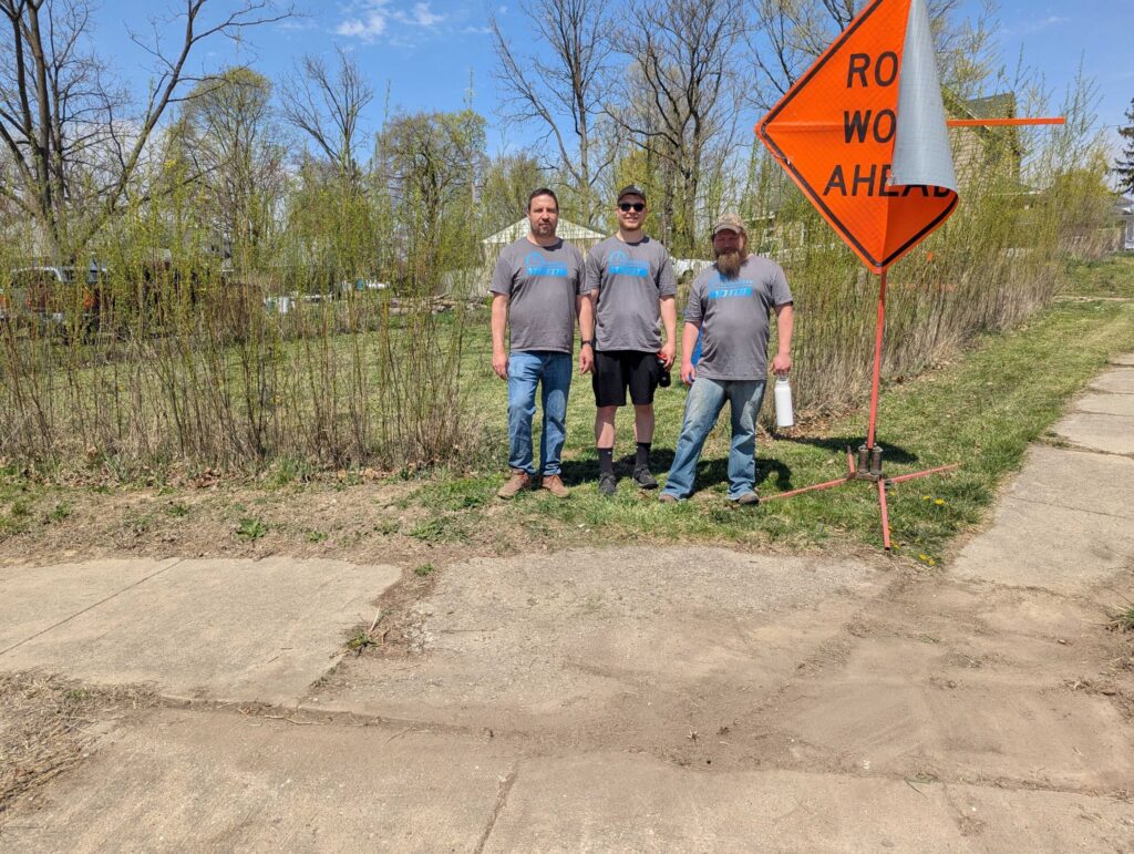 MOTOR volunteers in gray company t-shirts standing beside a ‘Road Work Ahead’ sign during a community service project on a sunny spring day.