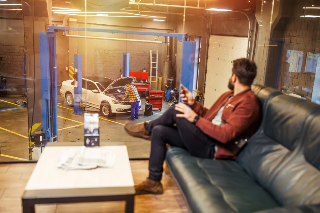 Customer in an auto shop waiting area watching a technician inspect a white sedan with its hood open inside a service bay.