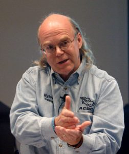 Instructor in a light blue training shirt gestures while explaining a concept, wearing glasses and speaking in an educational setting.