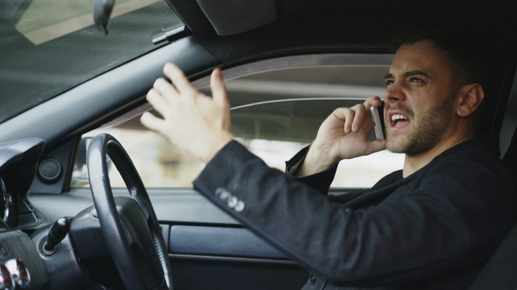 Driver sitting in a car, shouting in frustration while talking on a cellphone, illustrating distracted driving and unsafe road behavior.
