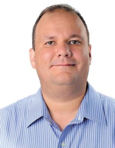 Studio headshot of a man with short dark hair, wearing a light blue striped button-down shirt, smiling slightly against a plain white background.