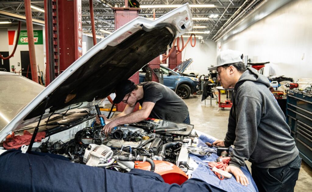 Two male mechanics working under the hood of a vehicle in an automotive repair garage.