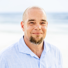 Professional headshot of a smiling man wearing a light blue shirt, standing outdoors near the beach with the ocean in the background.