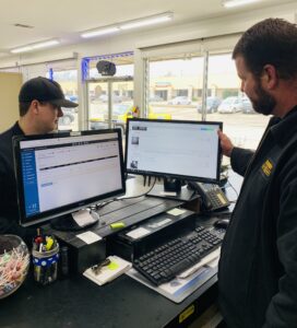 Automotive service professionals using MOTOR software on dual computer monitors at a repair shop counter to manage repair orders and customer information.