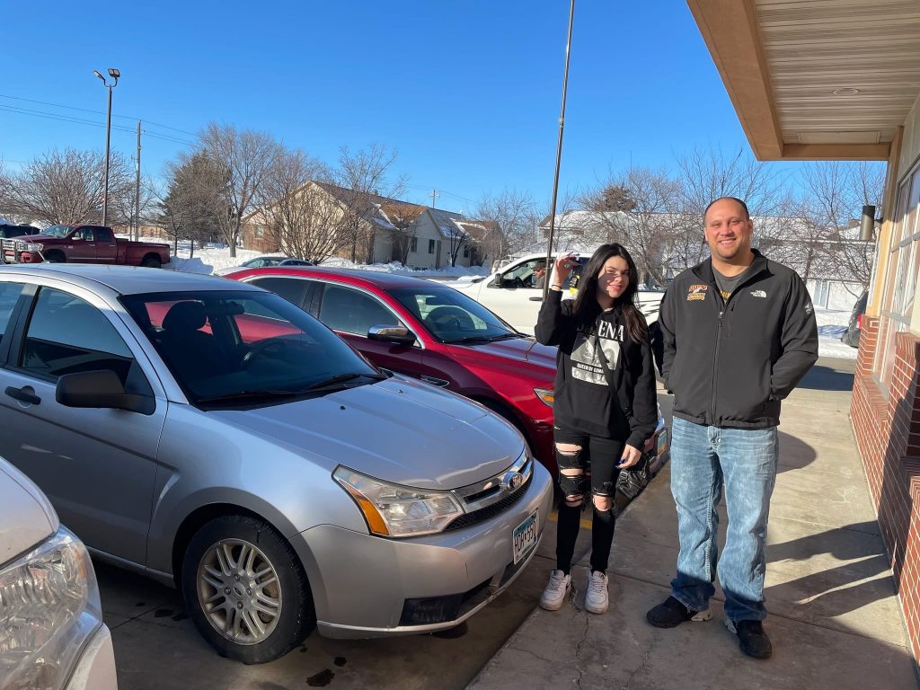 Automotive customer standing with a service professional outside a repair shop as she holds up her car keys beside a silver sedan and red vehicle on a sunny winter day.