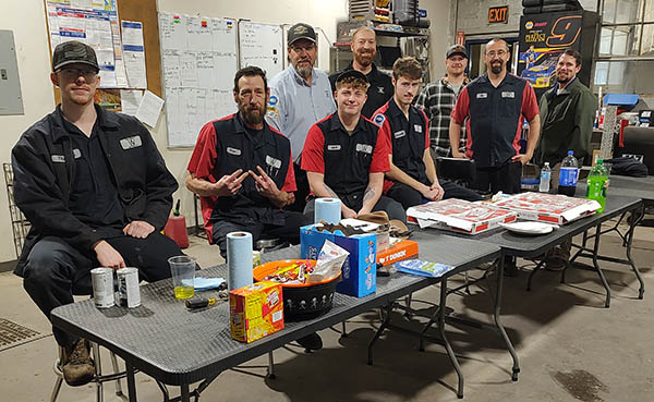 Automotive shop team gathered around tables with pizza and refreshments in a workshop, celebrating teamwork and employee camaraderie.