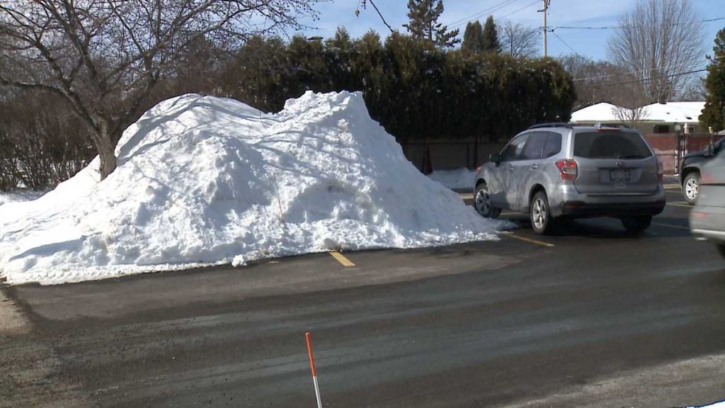 Huge mound of plowed snow in a parking lot beside a parked gray SUV after a winter snowstorm.