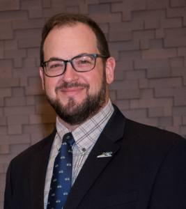 Professional headshot of a bearded man in glasses wearing a suit, patterned shirt and tie, standing against a textured wall at an industry event.