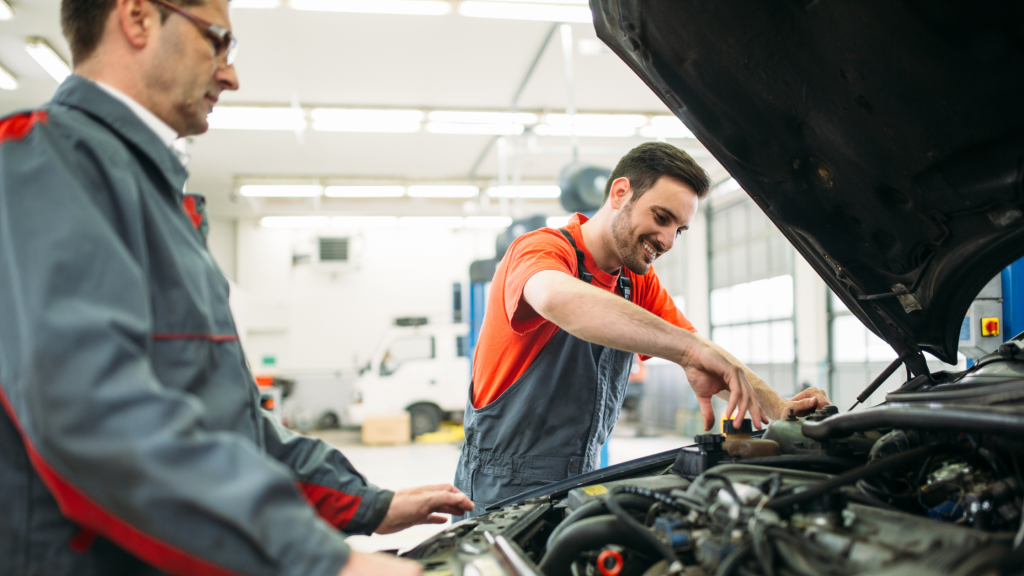 Auto mechanic smiling while repairing a car engine in a professional repair shop, with a colleague observing during vehicle maintenance.